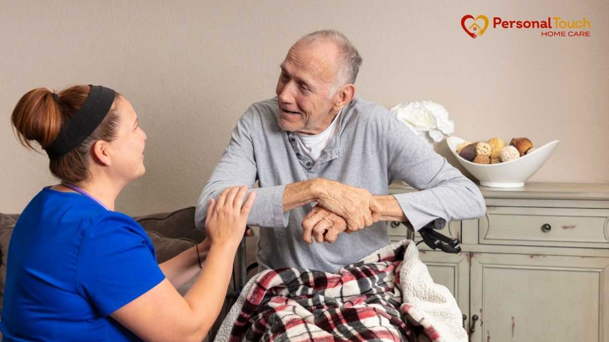 Caregiver in scrubs talking and holding hands with an elderly man in a wheelchair