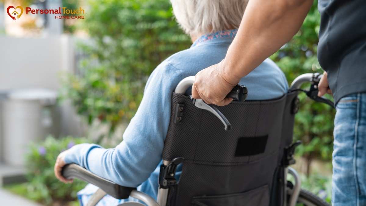 Caregiver pushing an elderly person in a wheelchair outdoors near a garden