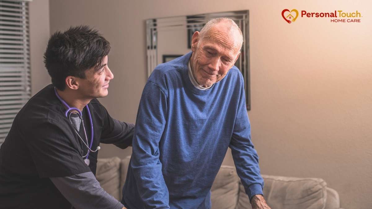 Nurse supporting an elderly man as he stands up from a couch at home